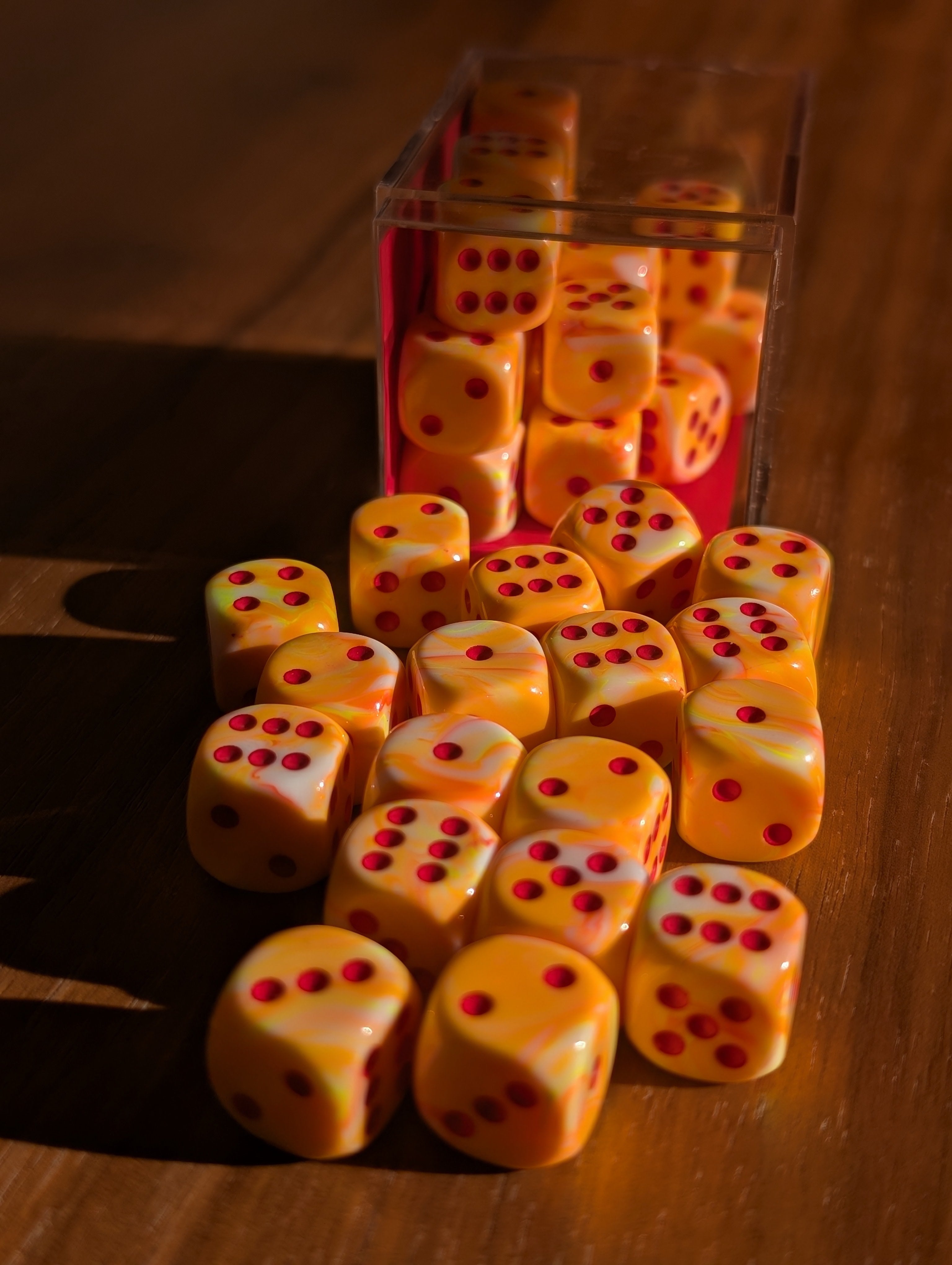 A set of 12mm six-sided dice spills out of a clear container toward the camera. They are neon orange swirled together with white highlights and dotted with red.
