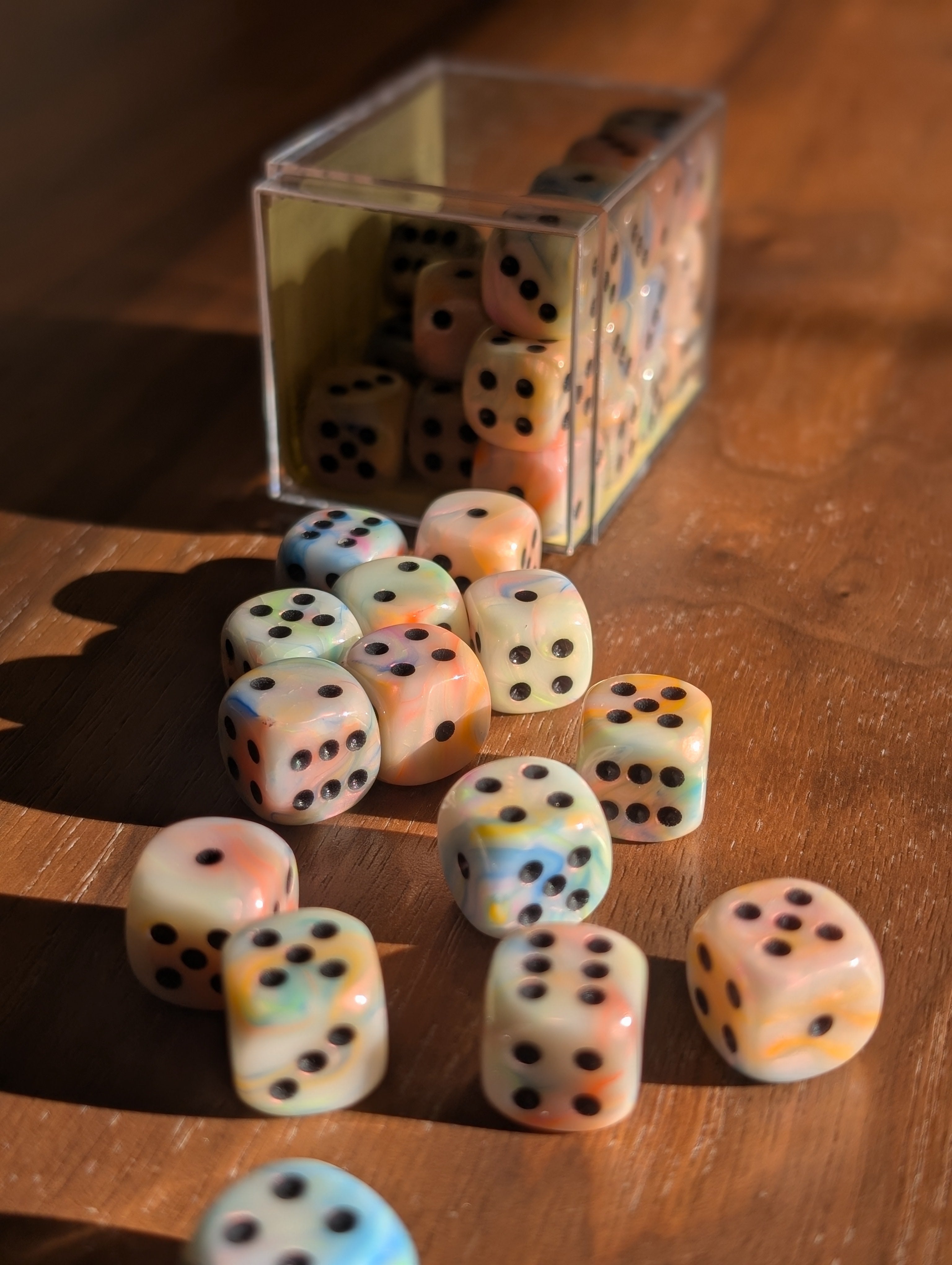 A set of 12mm dice spills out of a clear case toward the camera. They are several colours of marbled, pale resin with black dots.